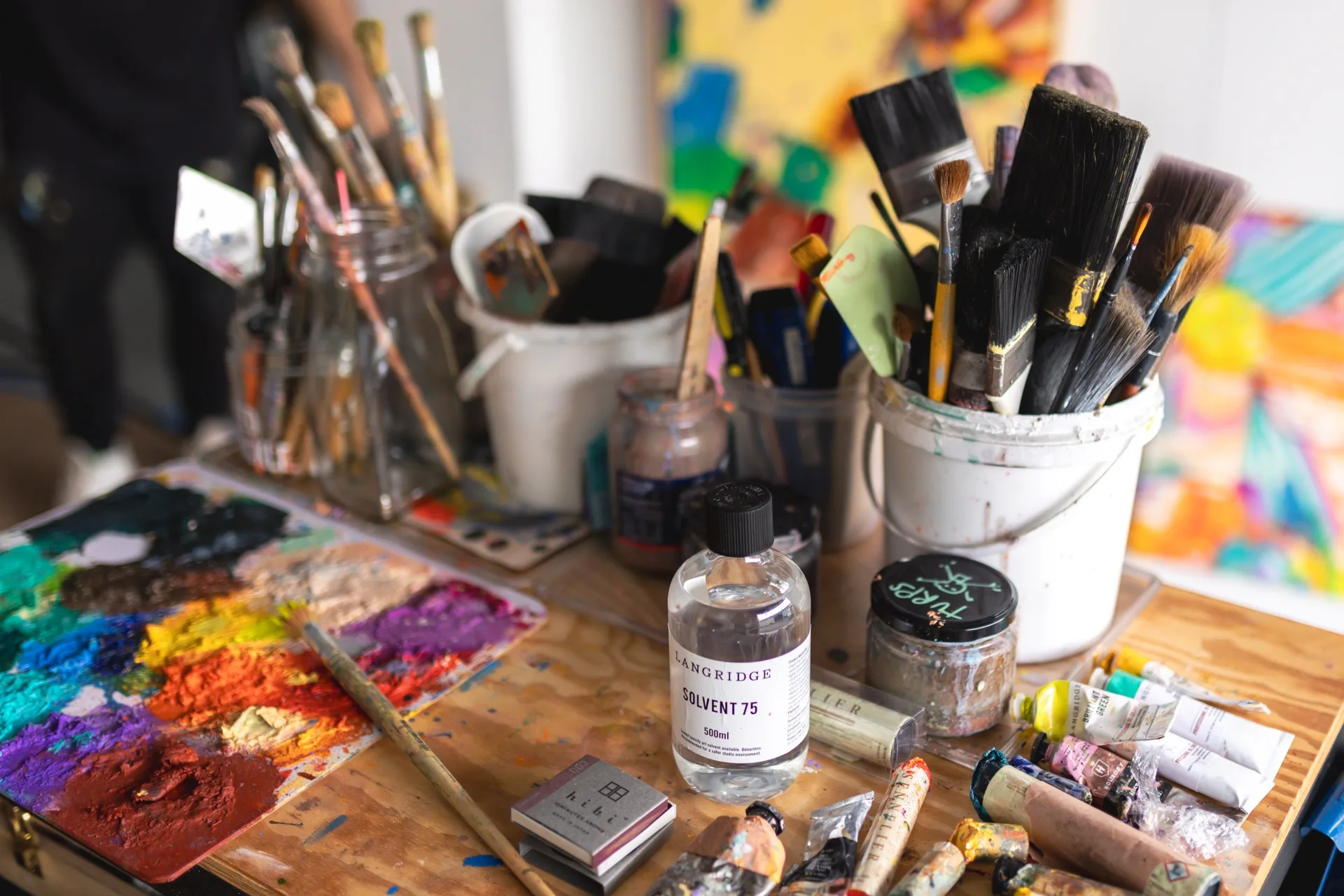 Close up photo of a painter's desk, including paint palette, tubes of oil paint, buckets of paintbrushes and solvent.