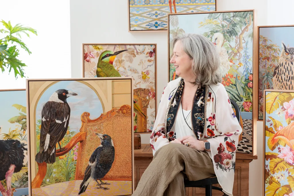 Woman sitting among vibrant bird paintings, smiling to the side, wearing a floral blouse with greenery in the background.