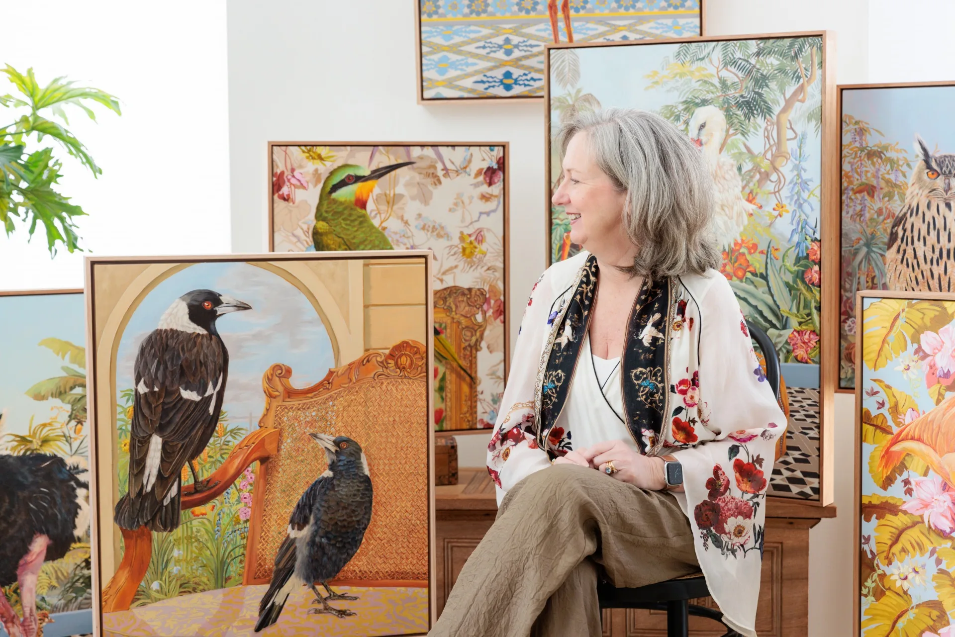 Woman sitting among vibrant bird paintings, smiling to the side, wearing a floral blouse with greenery in the background.