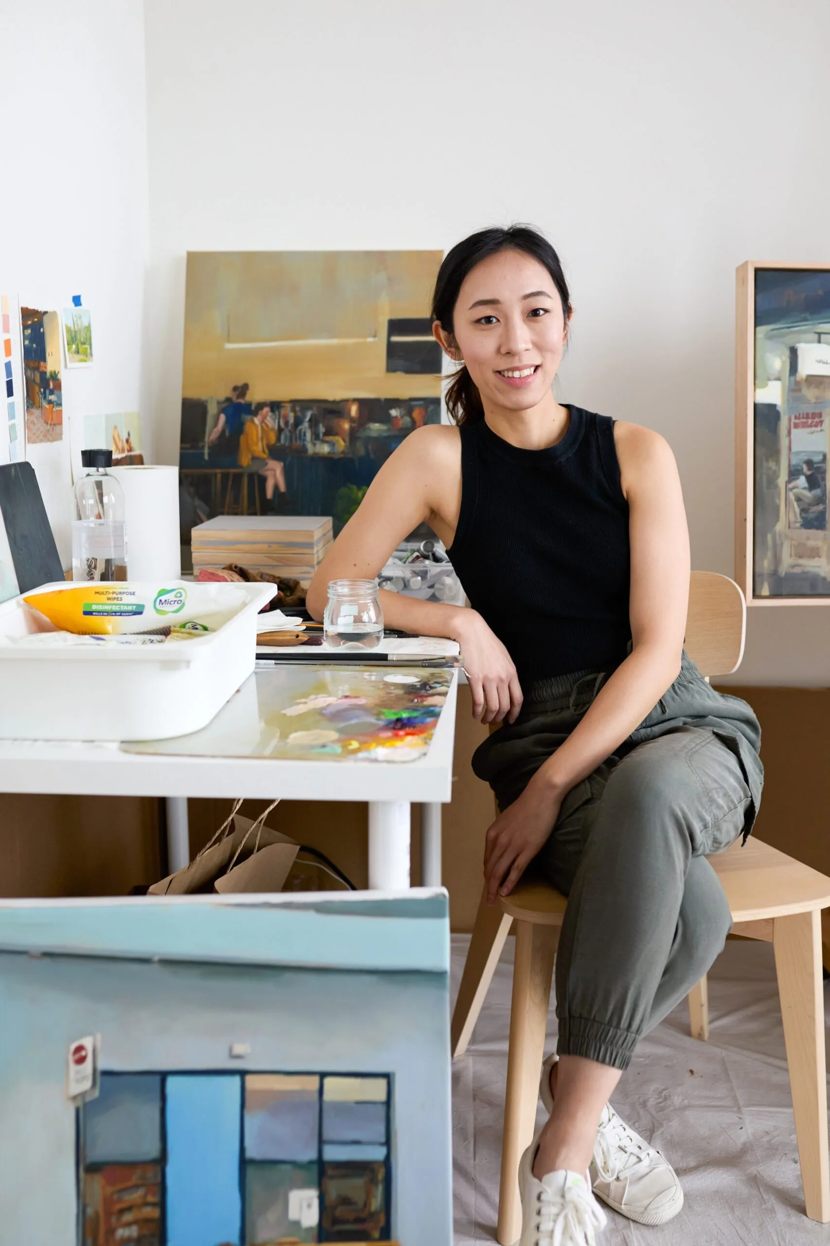 A woman sits in an art studio, surrounded by paintings and art supplies, smiling at the camera.