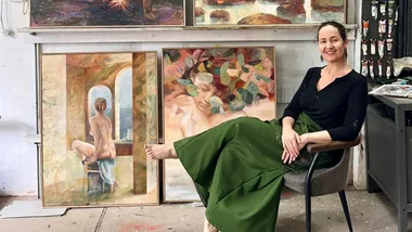 Artist sitting in a studio with several colorful paintings on shelves, smiling, wearing a green skirt and black top.