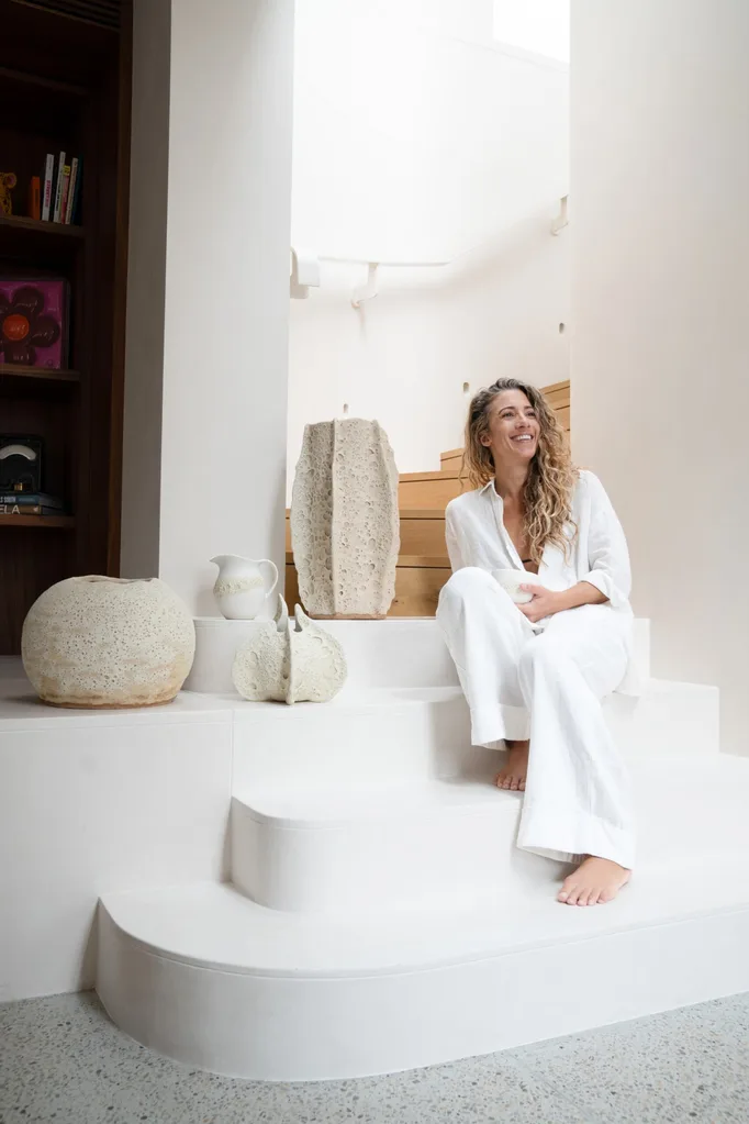 Person in white outfit sits on white stairs, surrounded by white ceramic art pieces in a bright room.