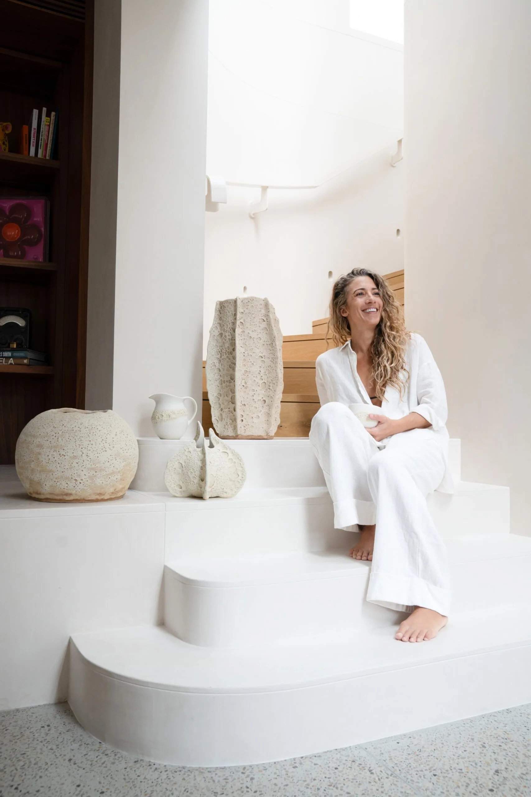 Person in white outfit sits on white stairs, surrounded by white ceramic art pieces in a bright room.