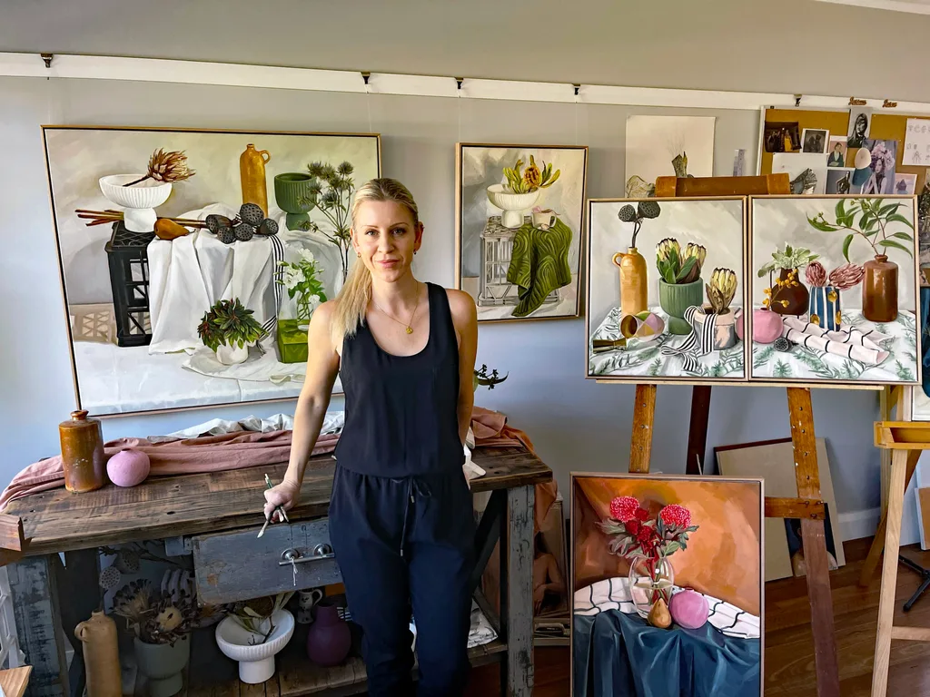 A woman in a studio stands next to a table and easels displaying detailed botanical still life paintings.