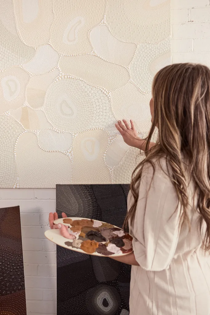 Woman in beige outfit standing in front of a white brick wall, holding a paint palette and looking at her painting, which is a contemporary Indigenous artwork.
