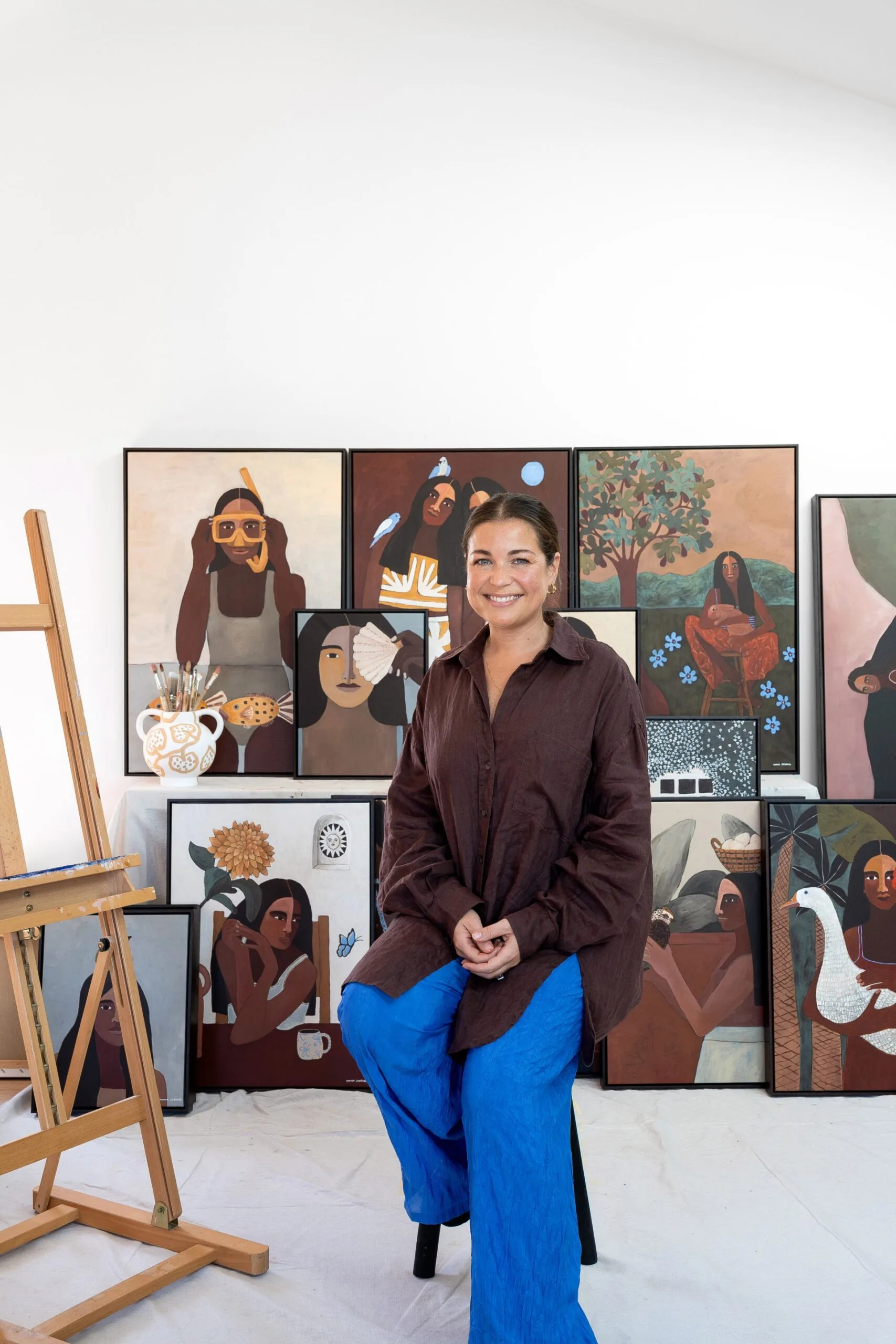 Artist sitting in a studio, surrounded by colorful paintings featuring women amongst nature.