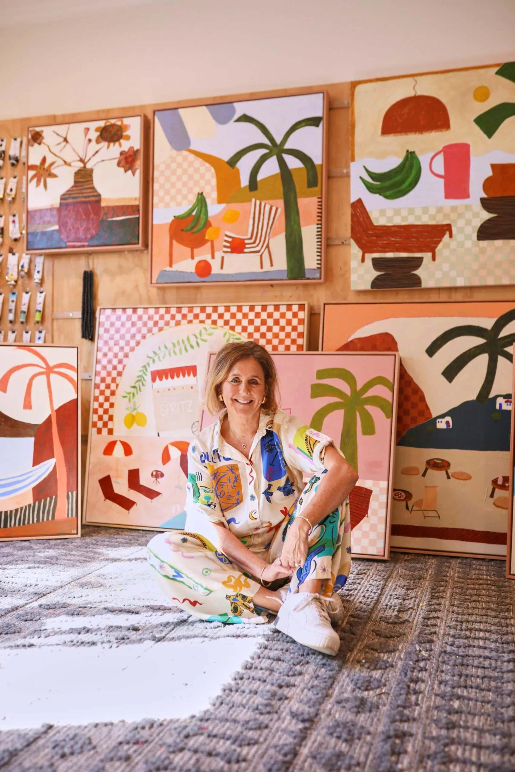 Artist sits on the floor in a colorful studio, surrounded by vibrant paintings of tropical themes.