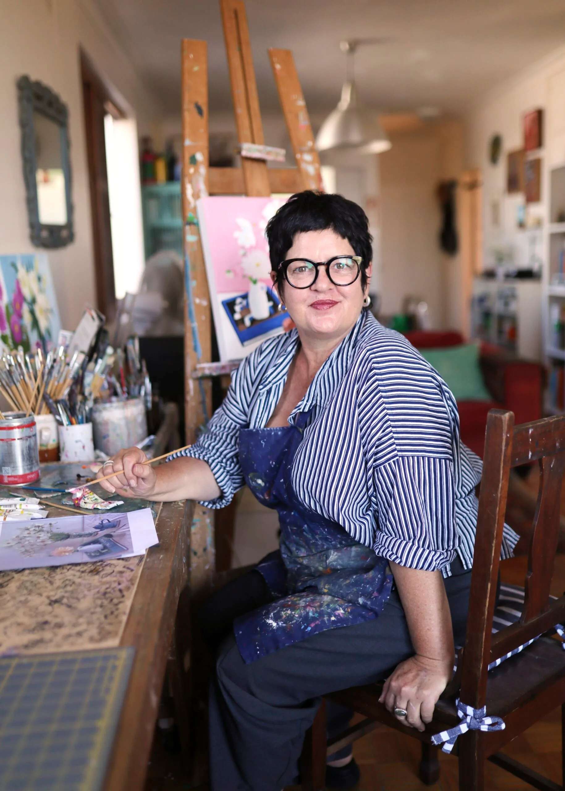 Artist in a striped shirt and apron sits in a studio, surrounded by paintbrushes and artworks on easels.