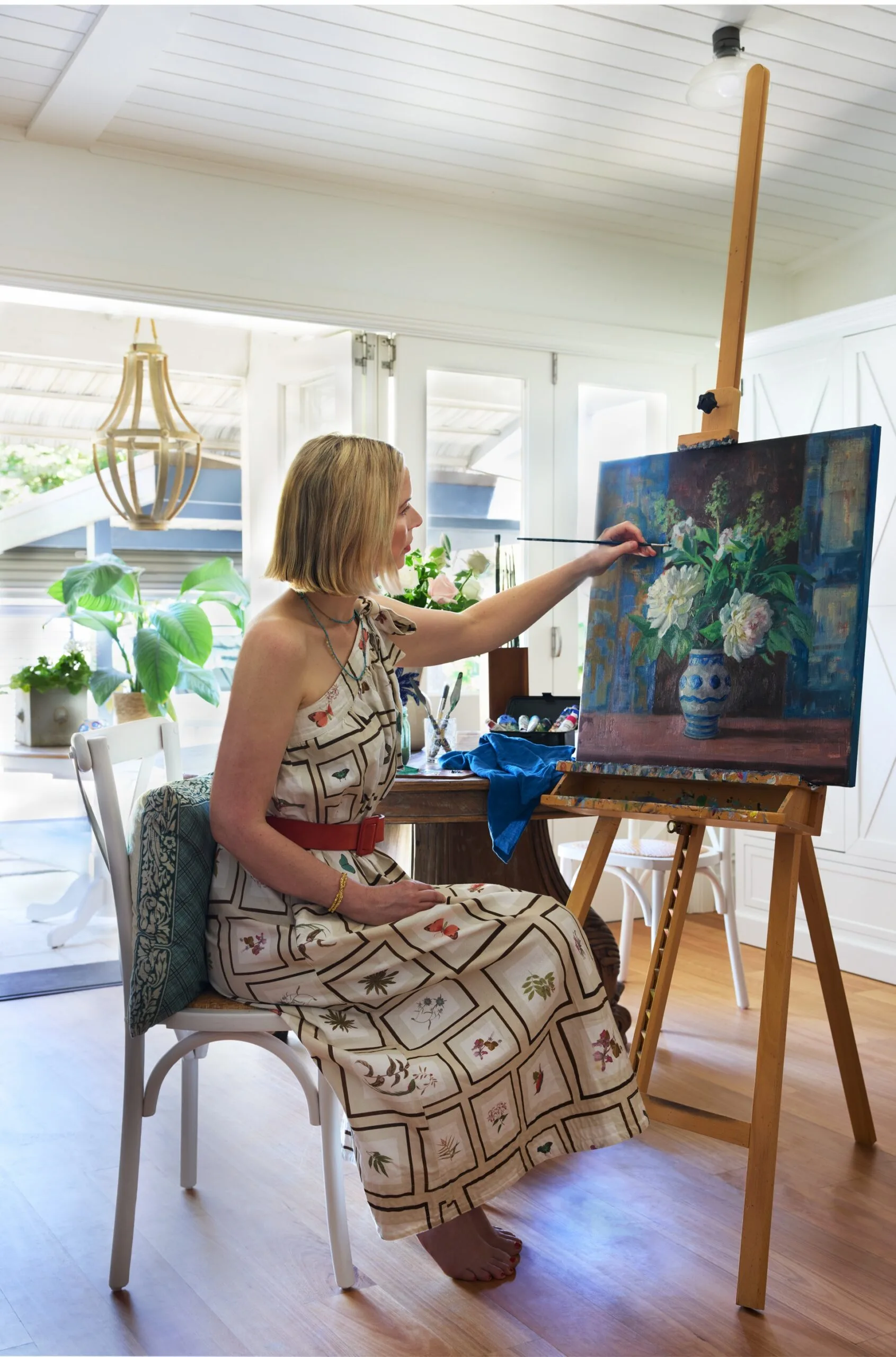 A woman in a patterned dress paints flowers on a canvas in a bright room with plants
