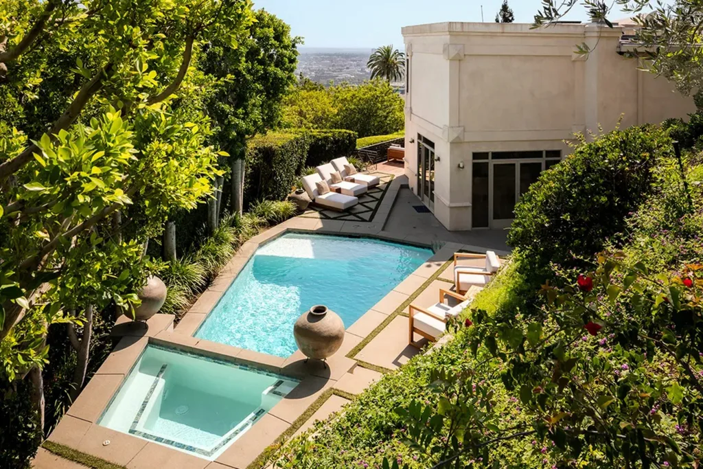 Aerial view of a luxurious backyard with a pool, hot tub, lounge chairs, lush greenery, and a city in the background.