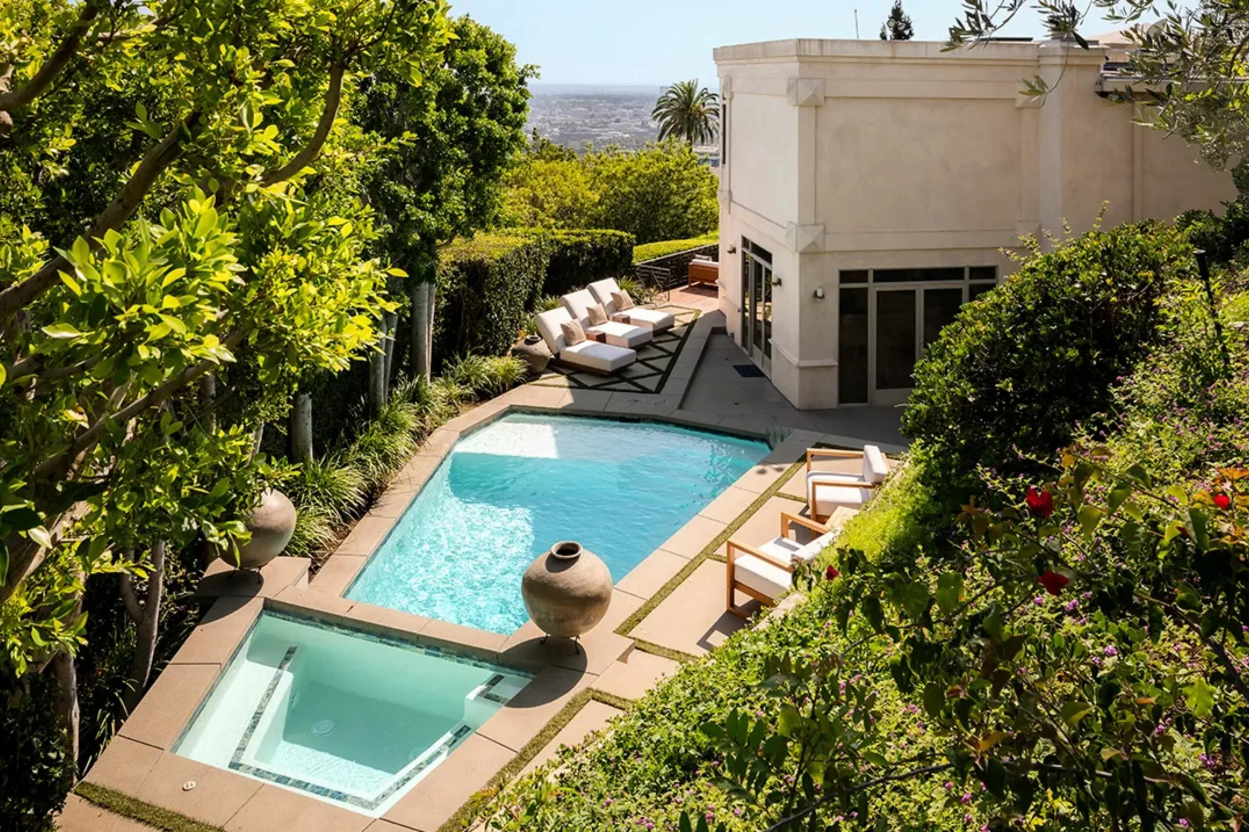 Aerial view of a luxurious backyard with a pool, hot tub, lounge chairs, lush greenery, and a city in the background.