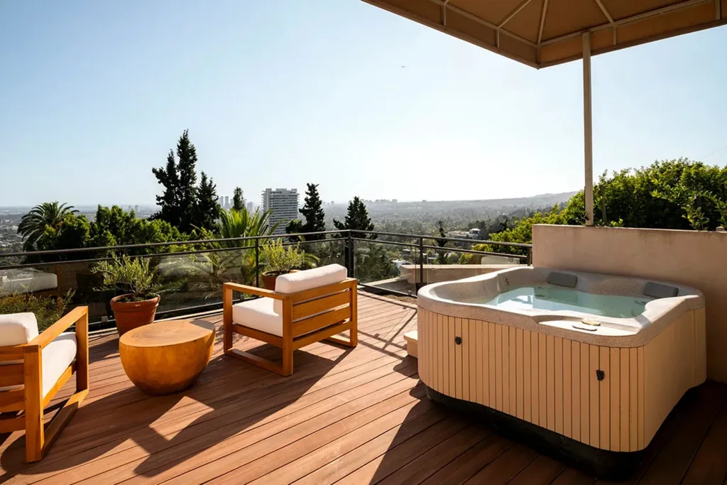 Rooftop deck with wooden chairs, a round table, and a hot tub, overlooking a city skyline under a clear sky.