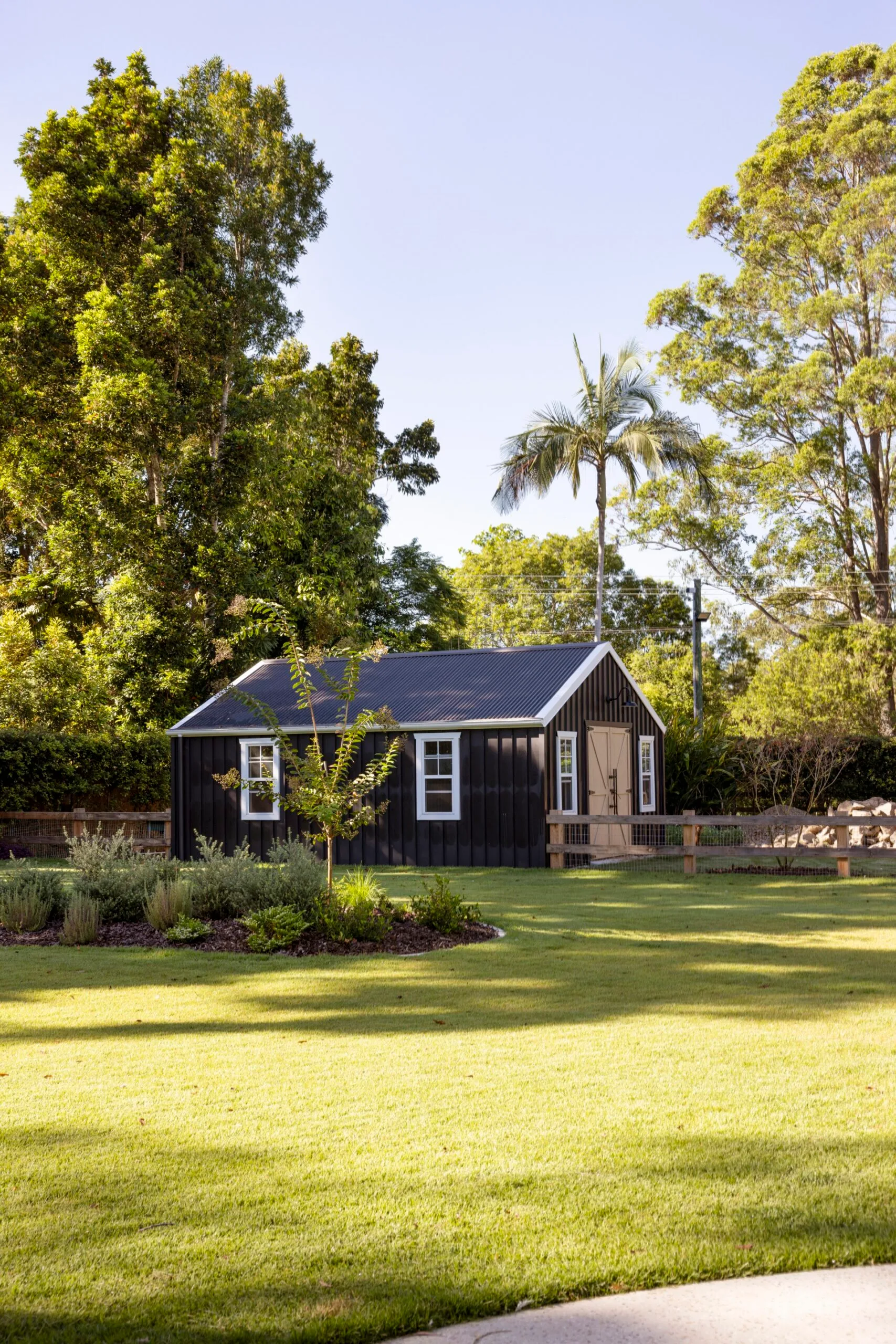Black shed with white trim in a sunny garden surrounded by trees and a neatly mowed lawn.