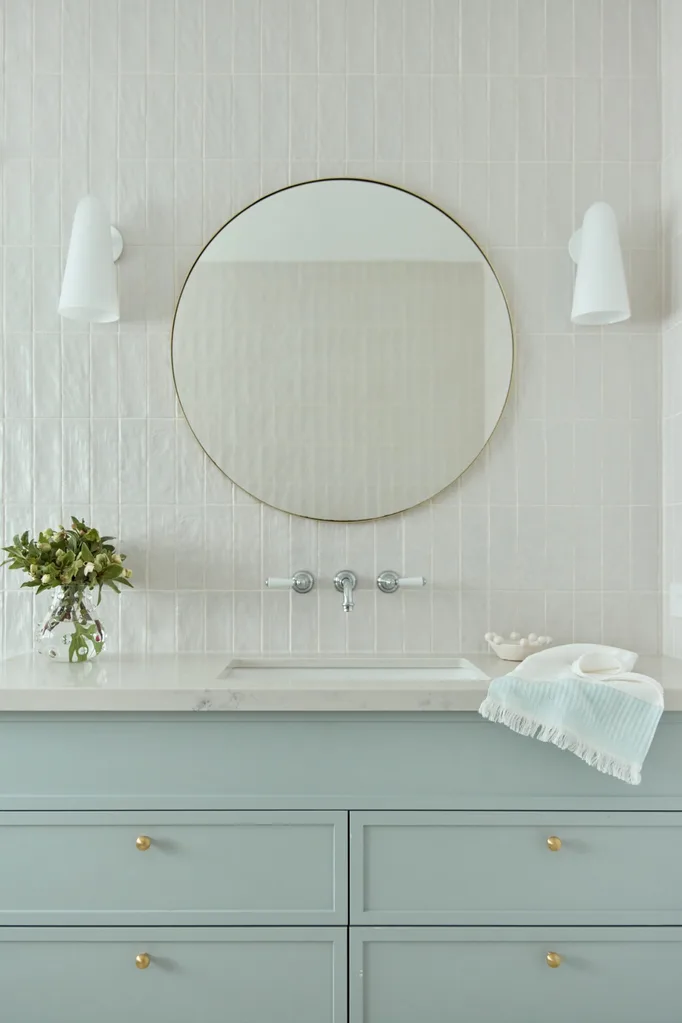 Modern bathroom with round mirror, light fixtures, green cabinetry, and decor items on countertop.