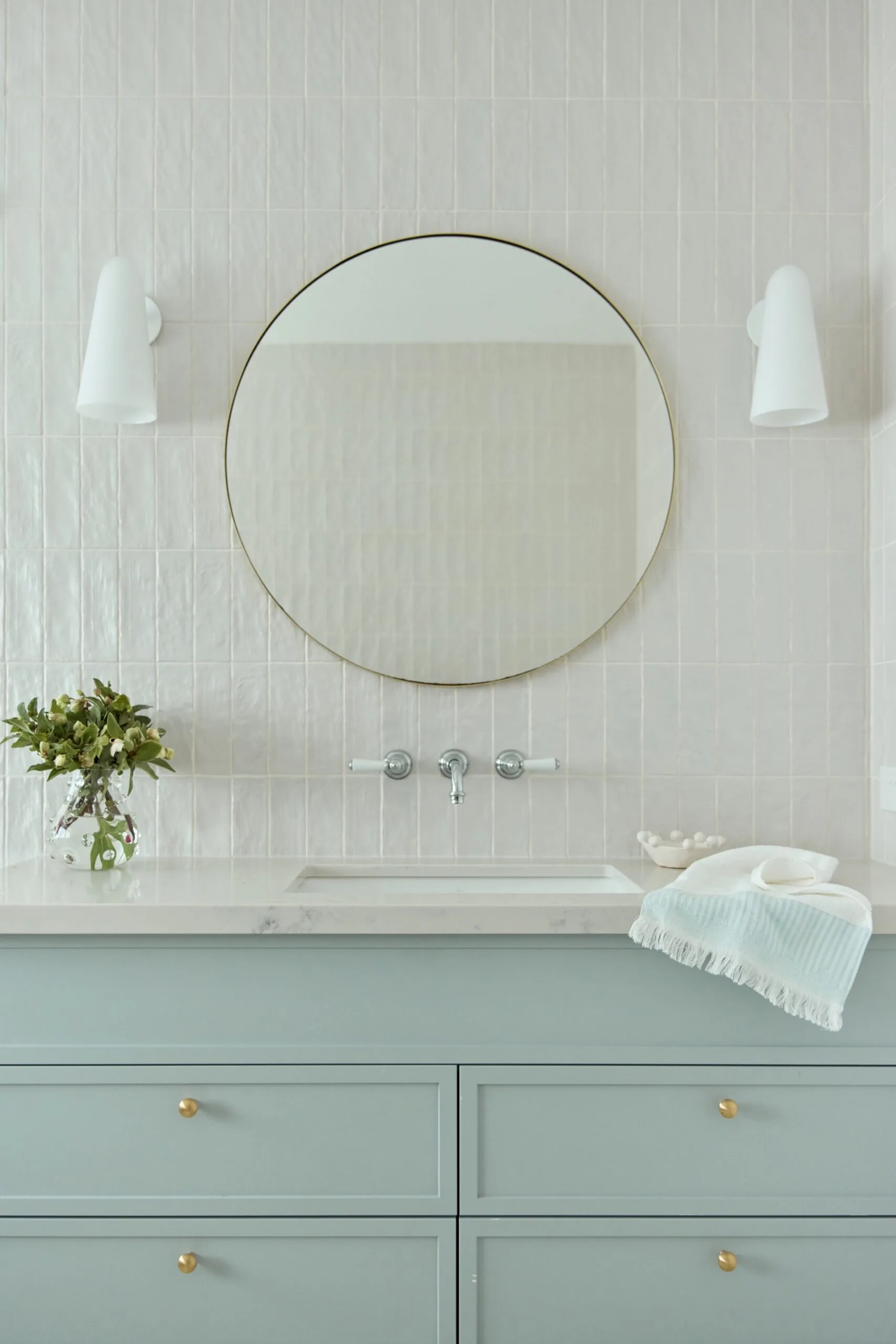 Modern bathroom with round mirror, light fixtures, green cabinetry, and decor items on countertop.