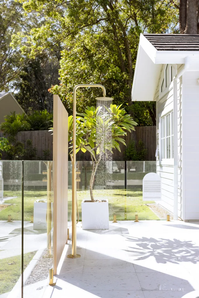 Outdoor shower next to a house with a glass fence and potted plants, set in a sunny garden.