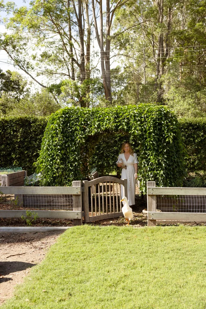 A woman in a white dress stands near a garden gate with lush greenery, looking at a duck walking on the path.