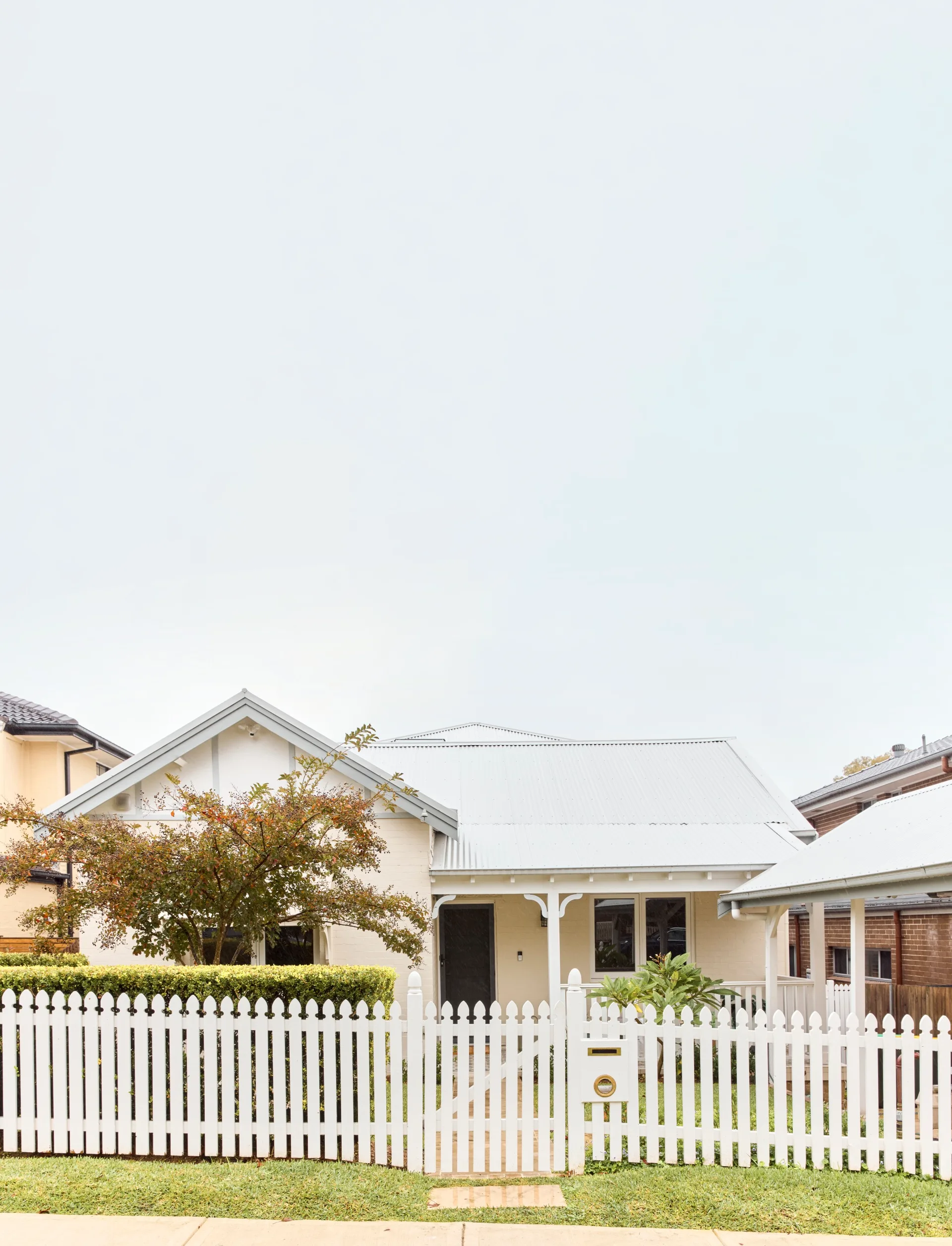 White picket-fenced house with a gabled roof and leafy garden under a cloudy sky.