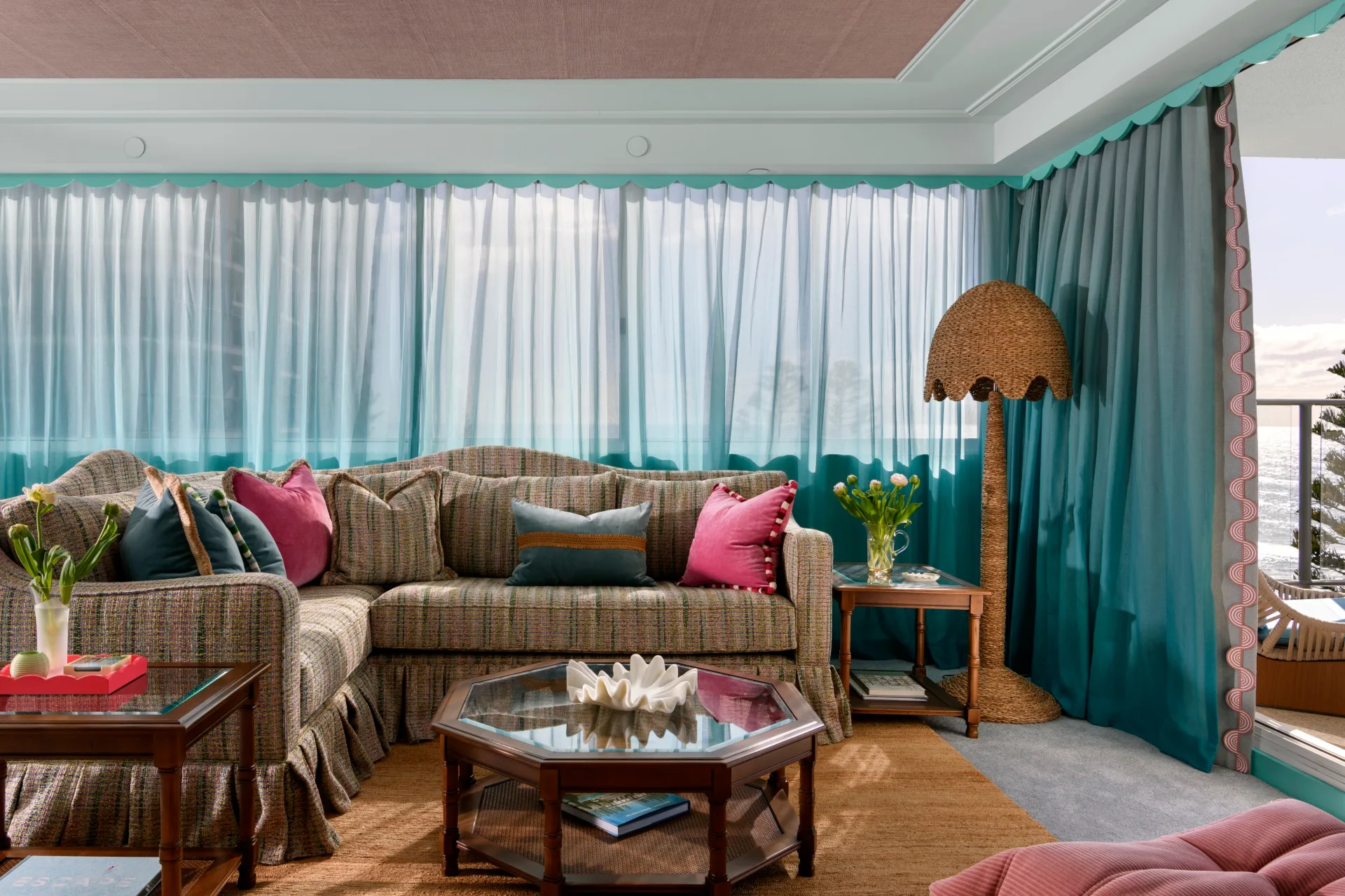 Cozy living room with corner sofa, colorful cushions, wicker lamp, teal curtains, and glass coffee table. Ocean view outside.