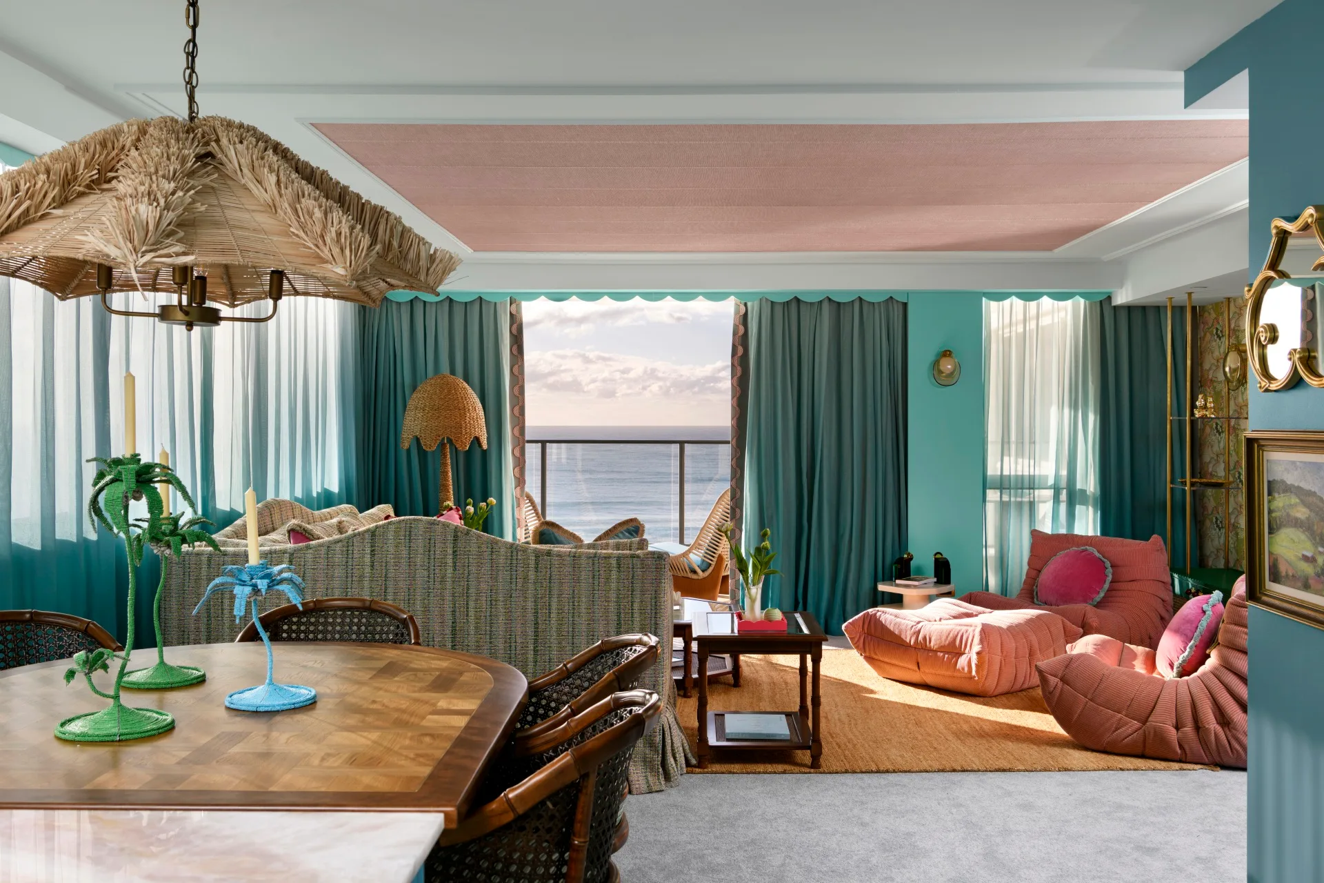 Colorful living room with retro furniture, ocean view, teal curtains, wooden dining table, and wicker chandelier.