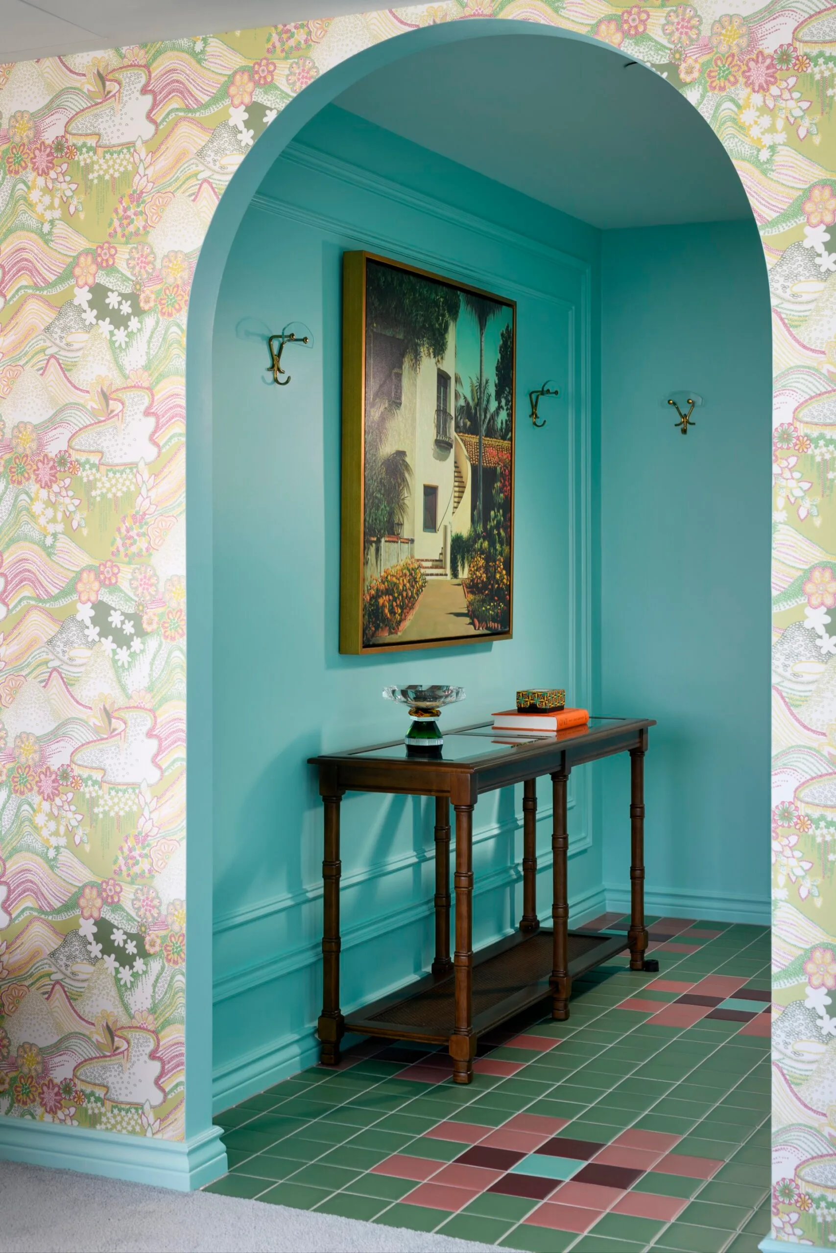 Arched hallway with floral wallpaper, teal walls, art, and a wooden console table on multicolored tile floor.