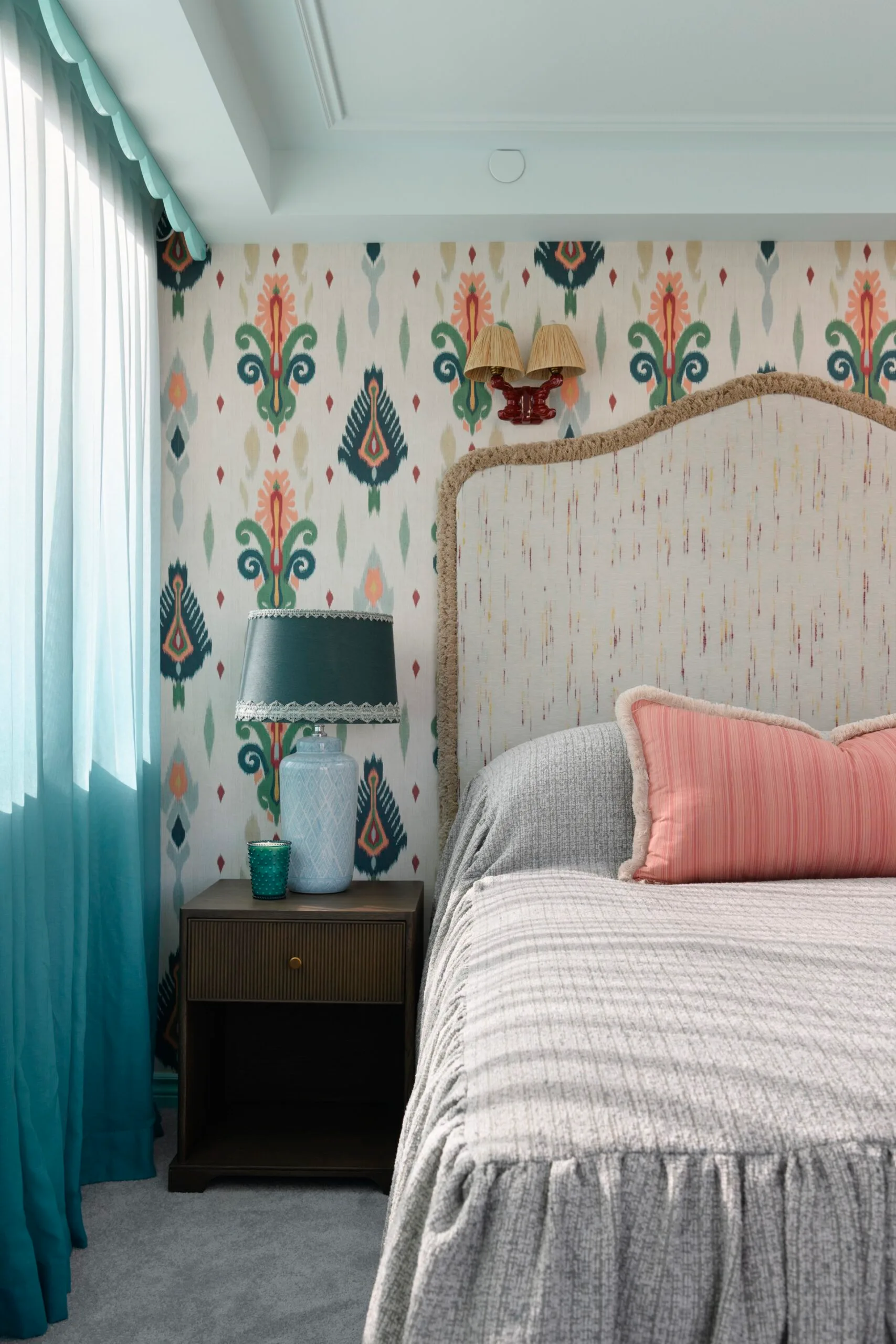 Cozy bedroom with patterned wallpaper, gray bedspread, red pillow, teal lamp, and sheer curtains.