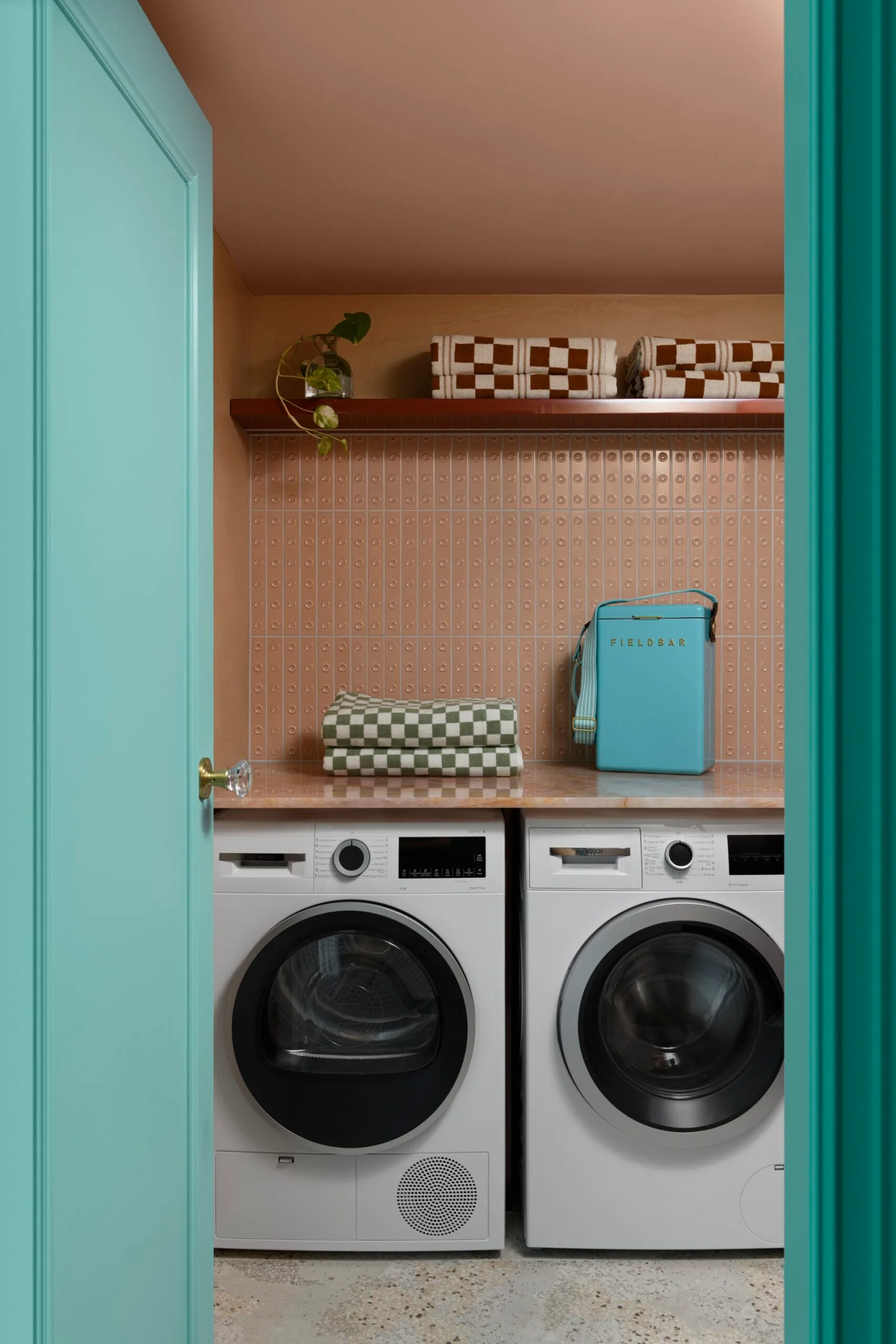 Turquoise laundry room with washer, dryer, patterned tiles, and a shelf holding towels, a plant, and a Fieldbar box.