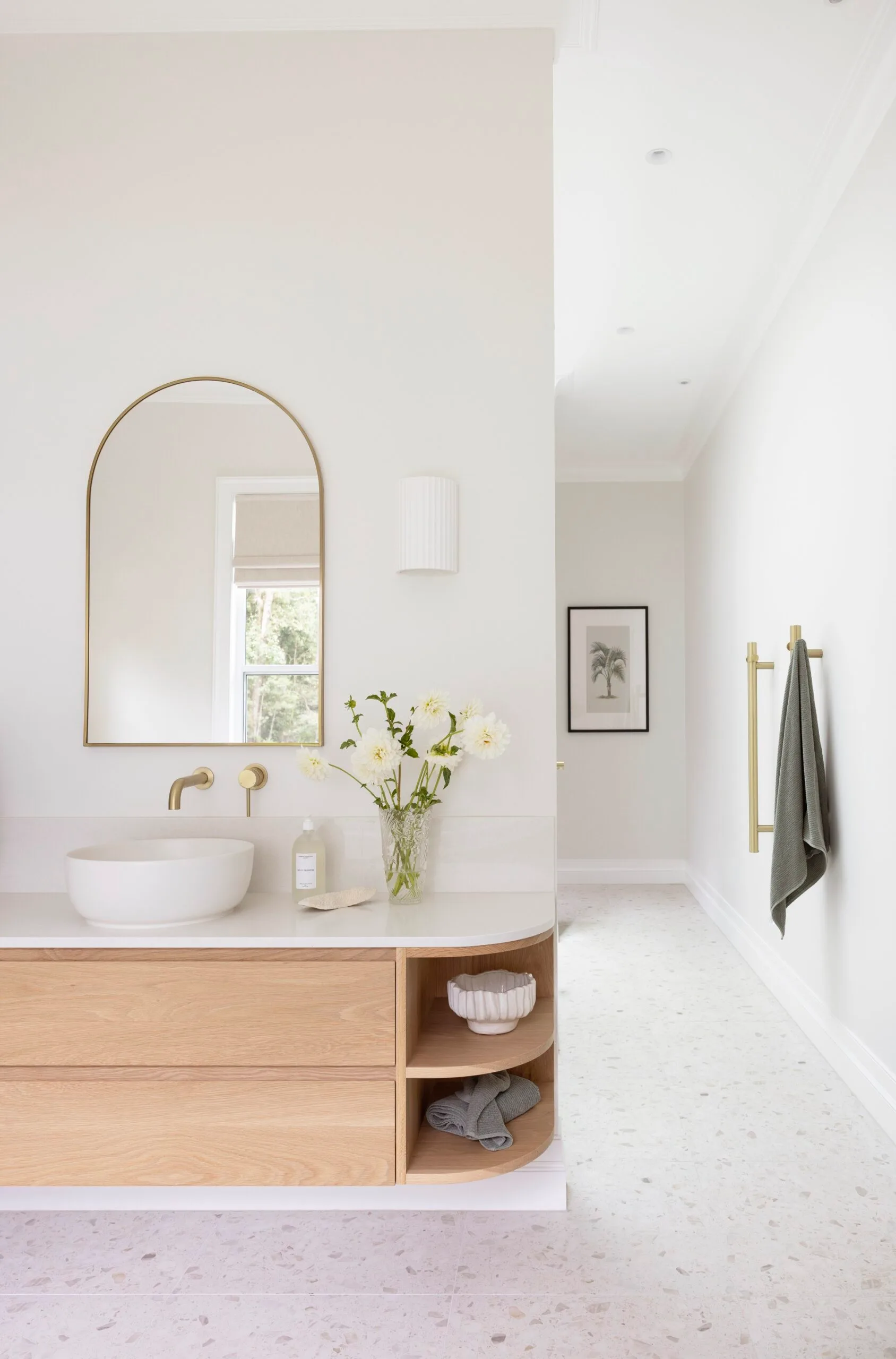 Modern bathroom with a round mirror, wooden vanity, white sink, flowers, and gold fixtures. Elegant, minimal design.