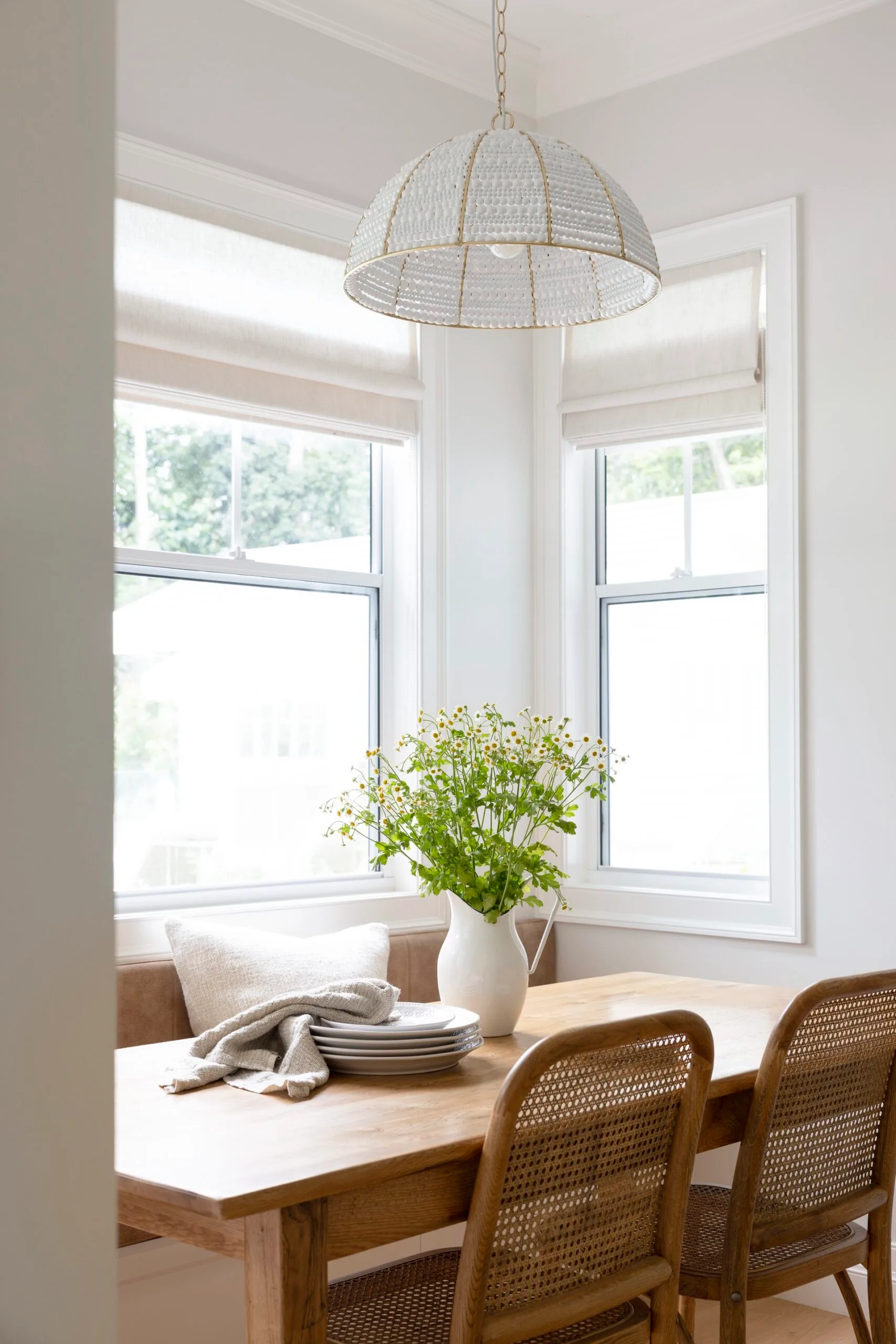 Cozy dining area with a wooden table, chairs, white jug with flowers, and soft light from a dome-shaped lamp.
