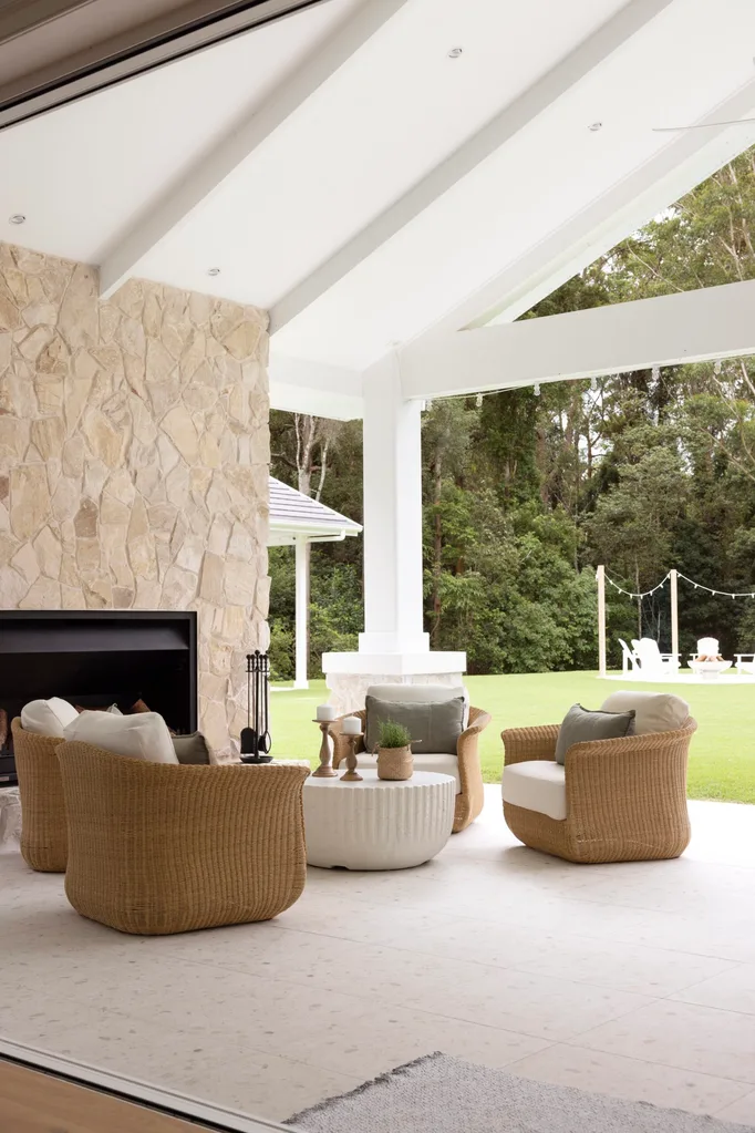 Outdoor patio with wicker chairs, stone fireplace, and a round white table with decor items, overlooking a green lawn.