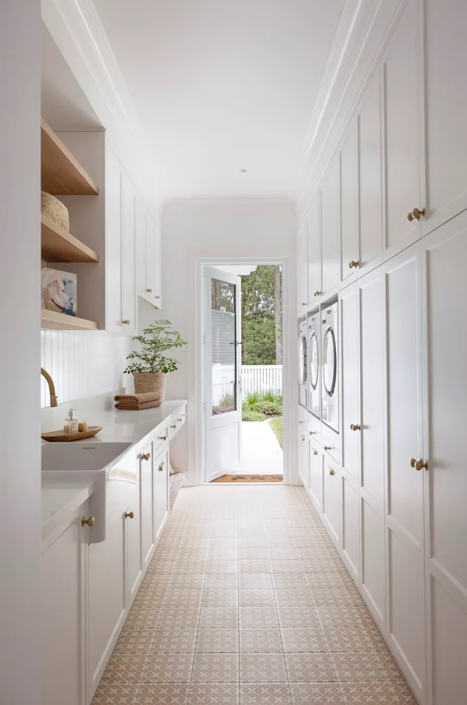 Bright laundry room with white cabinets, washer, dryer, farmhouse sink, plants, and open door to garden.