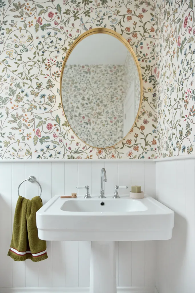 Floral wallpapered bathroom with oval mirror, white sink, green towel, and modern faucet.