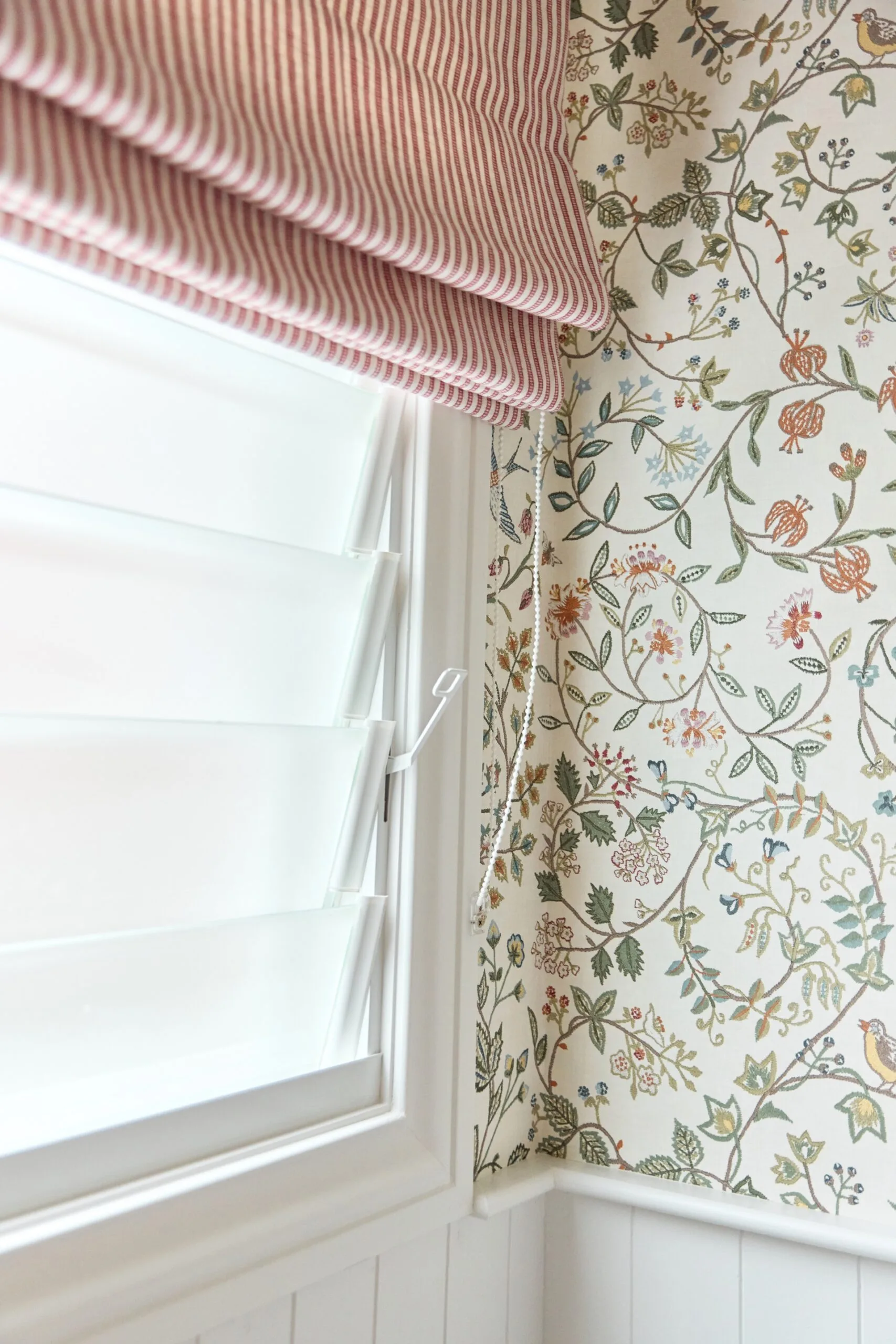 Red and white striped Roman shade next to floral-patterned wallpaper and white shutters.