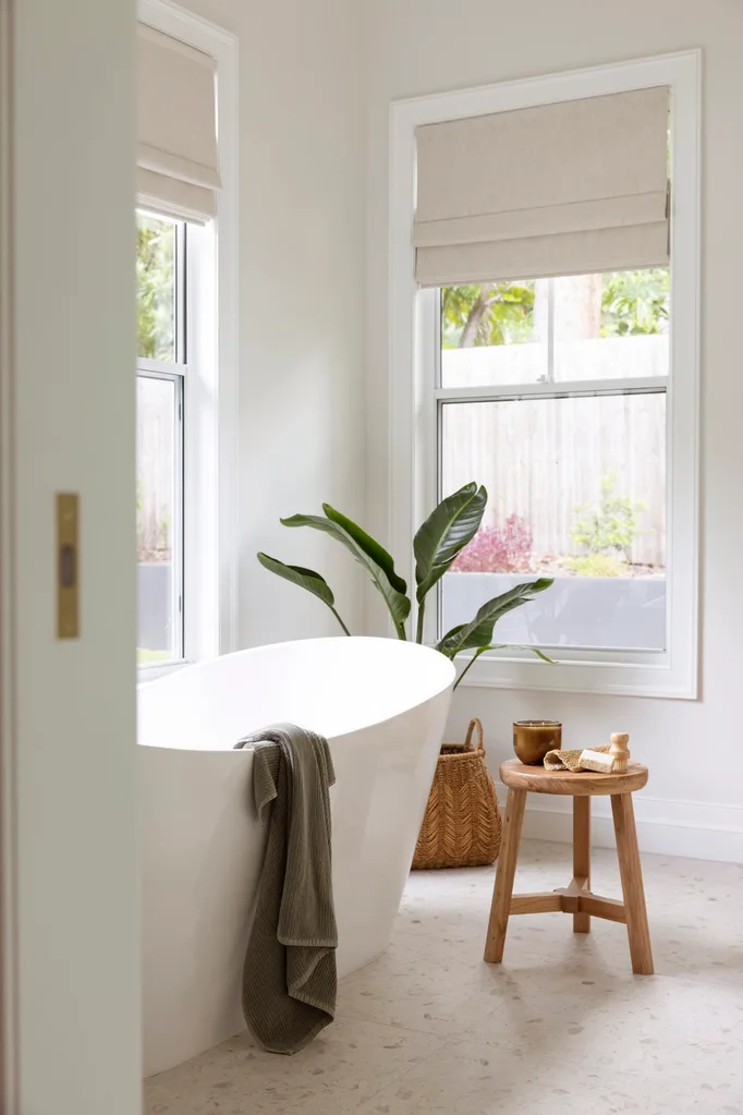 Bright bathroom with a white freestanding tub, a towel draped over it, and a wooden stool beside a large plant.