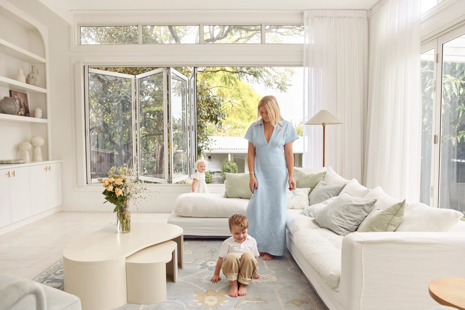 A woman in a light blue dress with two children in a bright, airy living room with an open window and cozy decor.