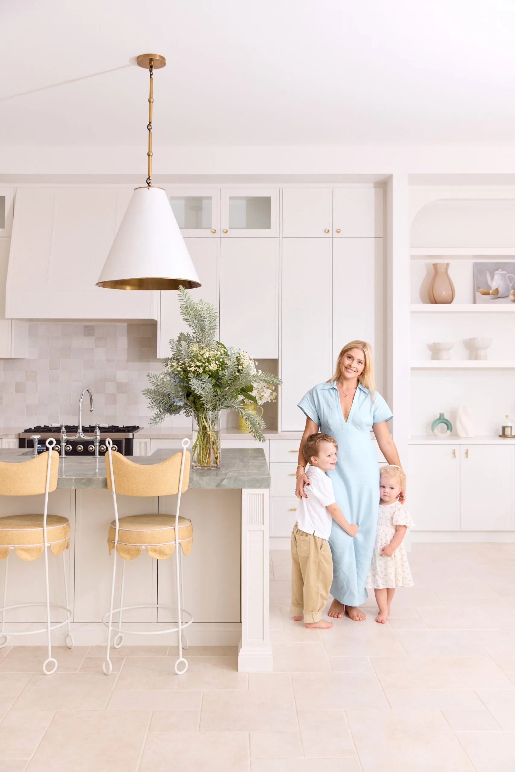 Woman in a blue dress with two children in a bright, modern kitchen with light decor and a bouquet on the island.