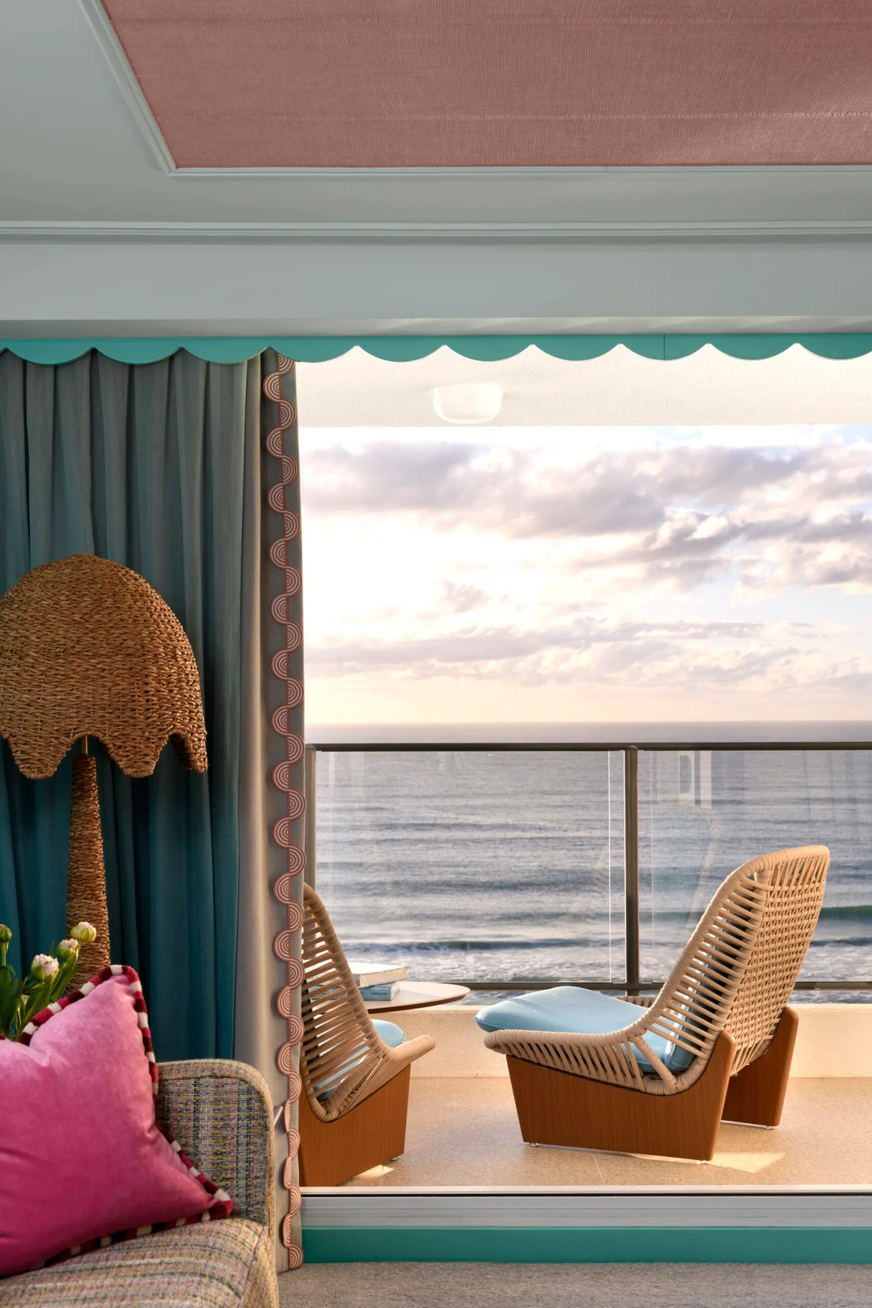 Interior with wicker chair, ocean view, pink pillow on sofa, and woven lamp; overlooking the sea through open balcony doors.