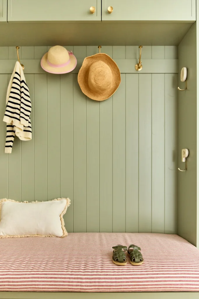 Green mudroom with striped bench, pillow, sandals, and hooks holding a sweater and two hats.
