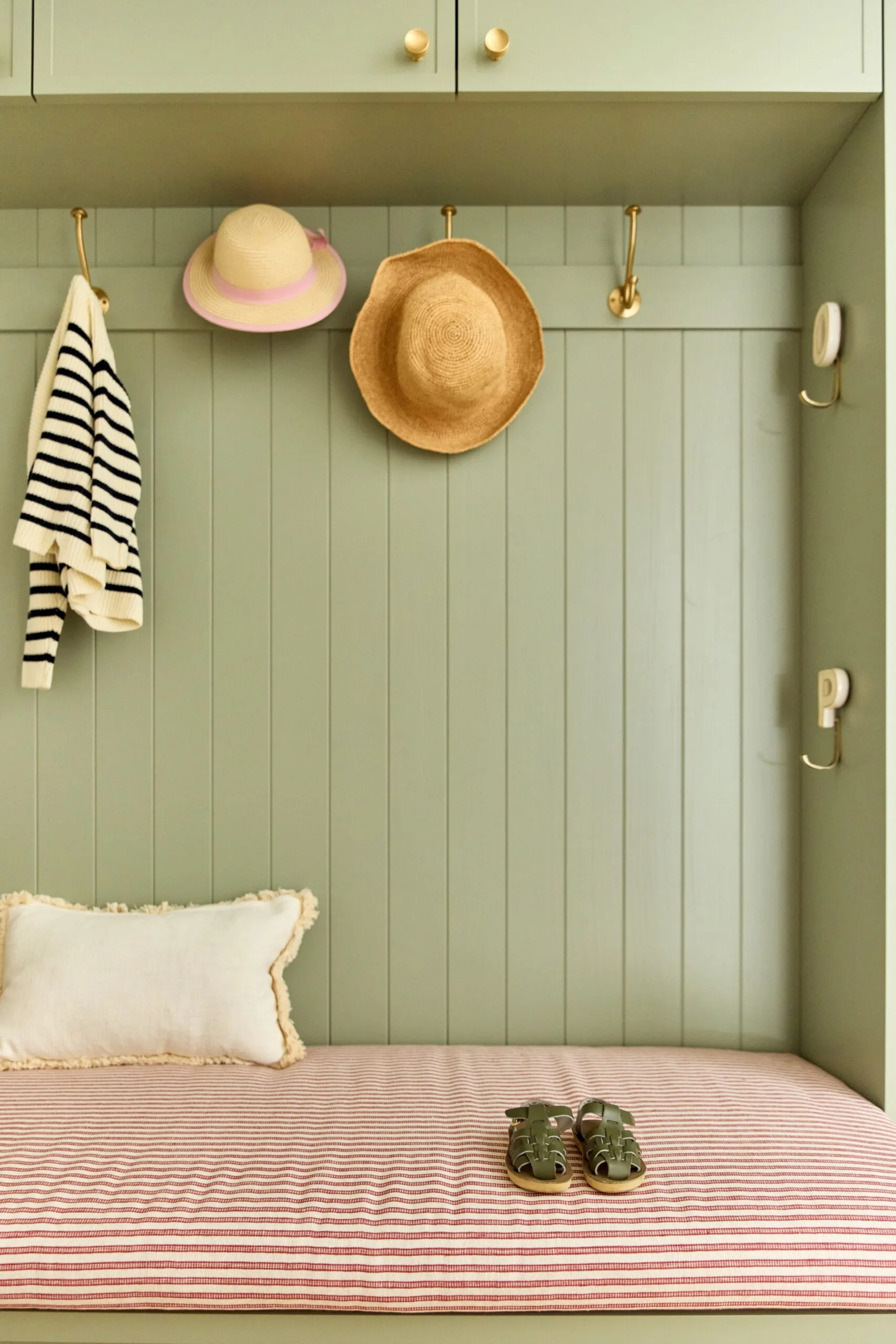 Green mudroom with striped bench, pillow, sandals, and hooks holding a sweater and two hats.