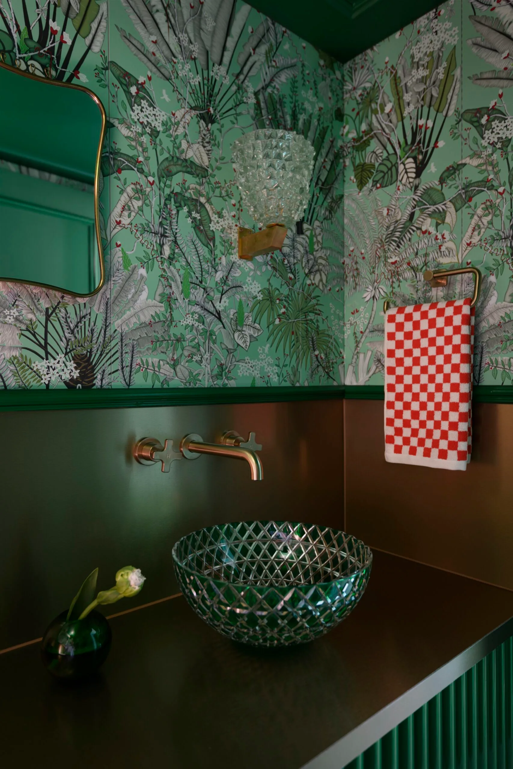 Bathroom with green floral wallpaper, glass bowl sink, brass faucet, and red-checkered towel.