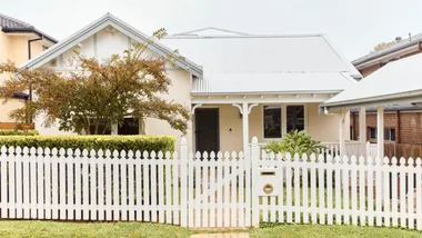 White picket-fenced house with a gabled roof and leafy garden under a cloudy sky.