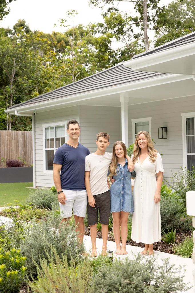 A family of four standing in front of a gray house with a garden, smiling at the camera.