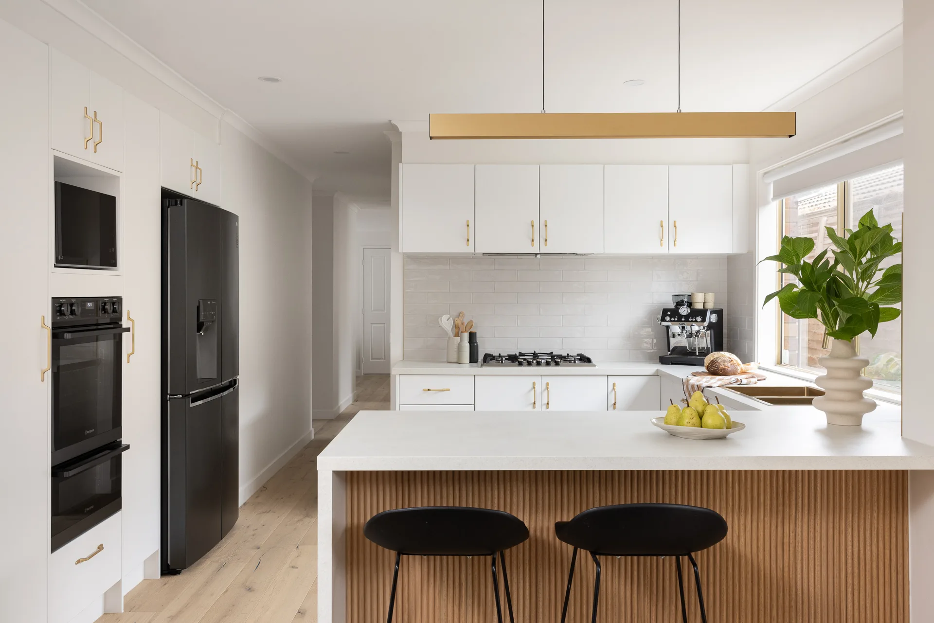 Modern kitchen with white cabinets, black appliances, and a kitchen island with black stools. A vase with green leaves and a bowl of pears sit on the island.
