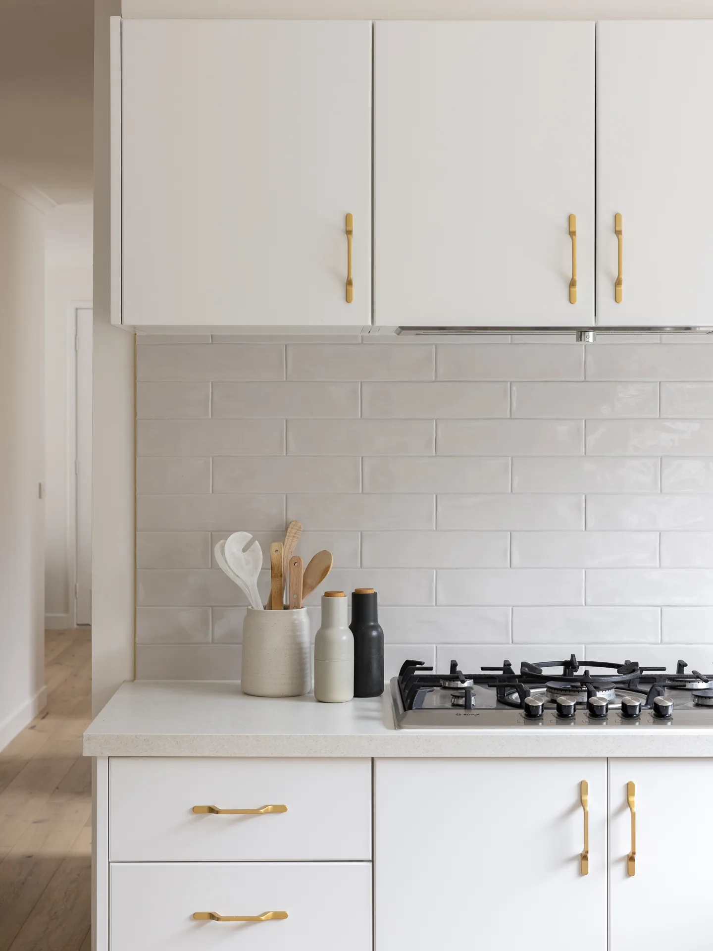 Modern kitchen with white cabinets, gold handles, gray backsplash, and a countertop with utensils and gas stove.