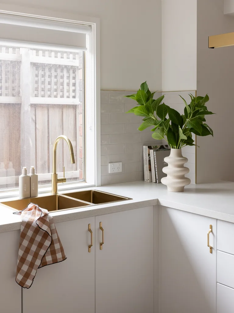 Modern kitchen corner with gold sink, white cabinets, potted plant in a white vase, and checkered towel on counter.