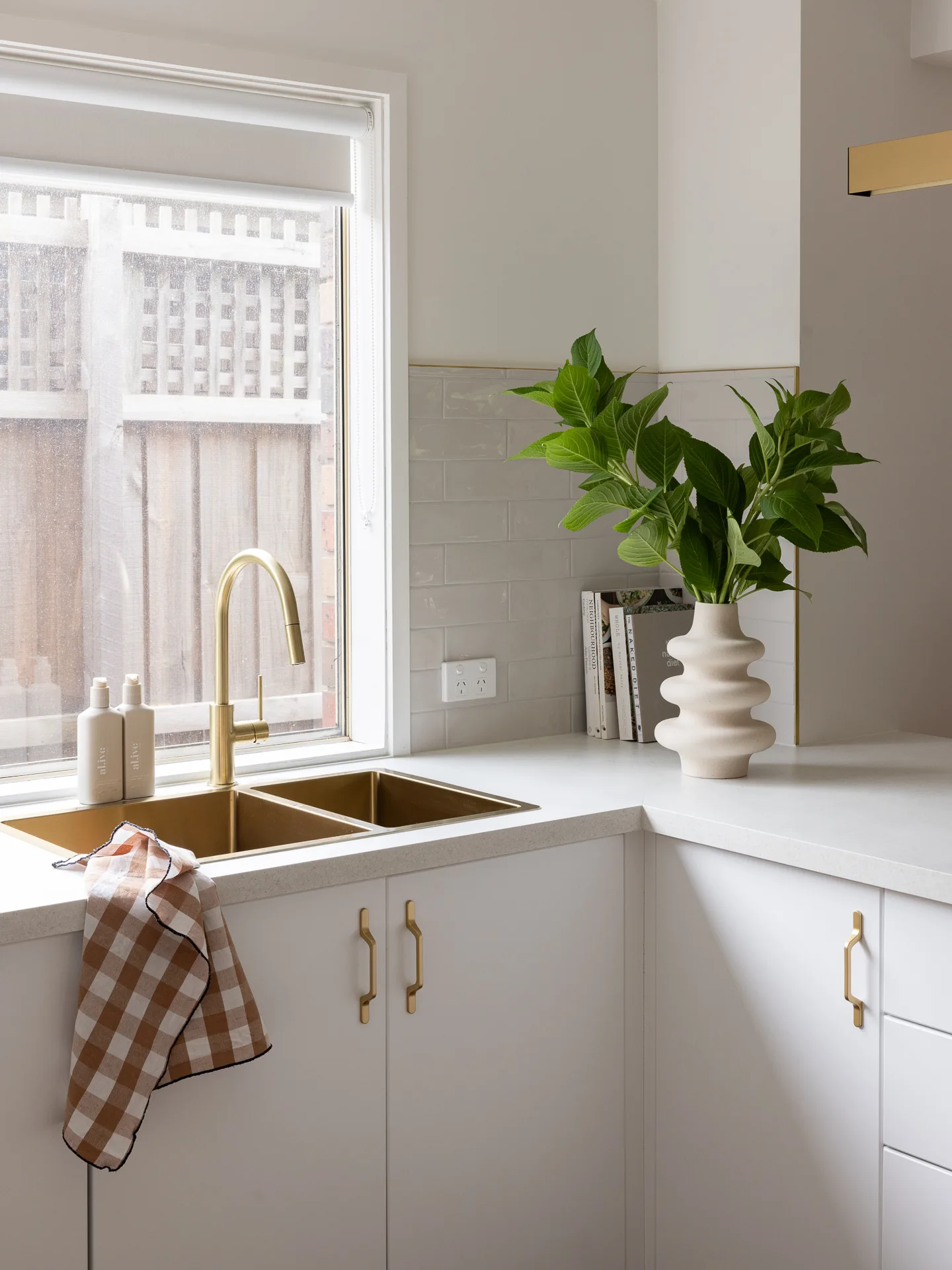 Modern kitchen corner with gold sink, white cabinets, potted plant in a white vase, and checkered towel on counter.