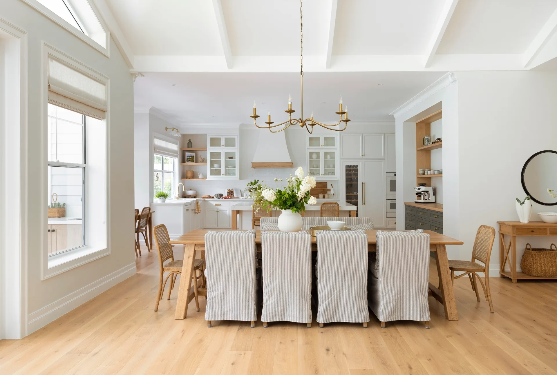 Bright kitchen and dining room with white walls, wooden floors, a large table with chairs, and decorative plants.
