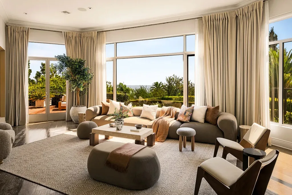 Spacious living room with large windows, beige sofas, plants, and a view of a sunny outdoor terrace. Dolly Parton's Hollywood Hills home.