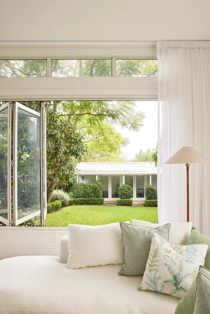 Bright living room with an open window view of a lush garden, featuring a white sofa with green and patterned pillows.