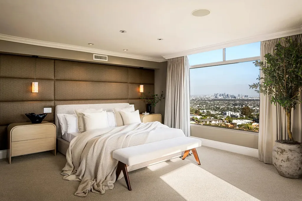 Modern bedroom with beige tones, large window showing city view, comfortable bed, and decorative plants.